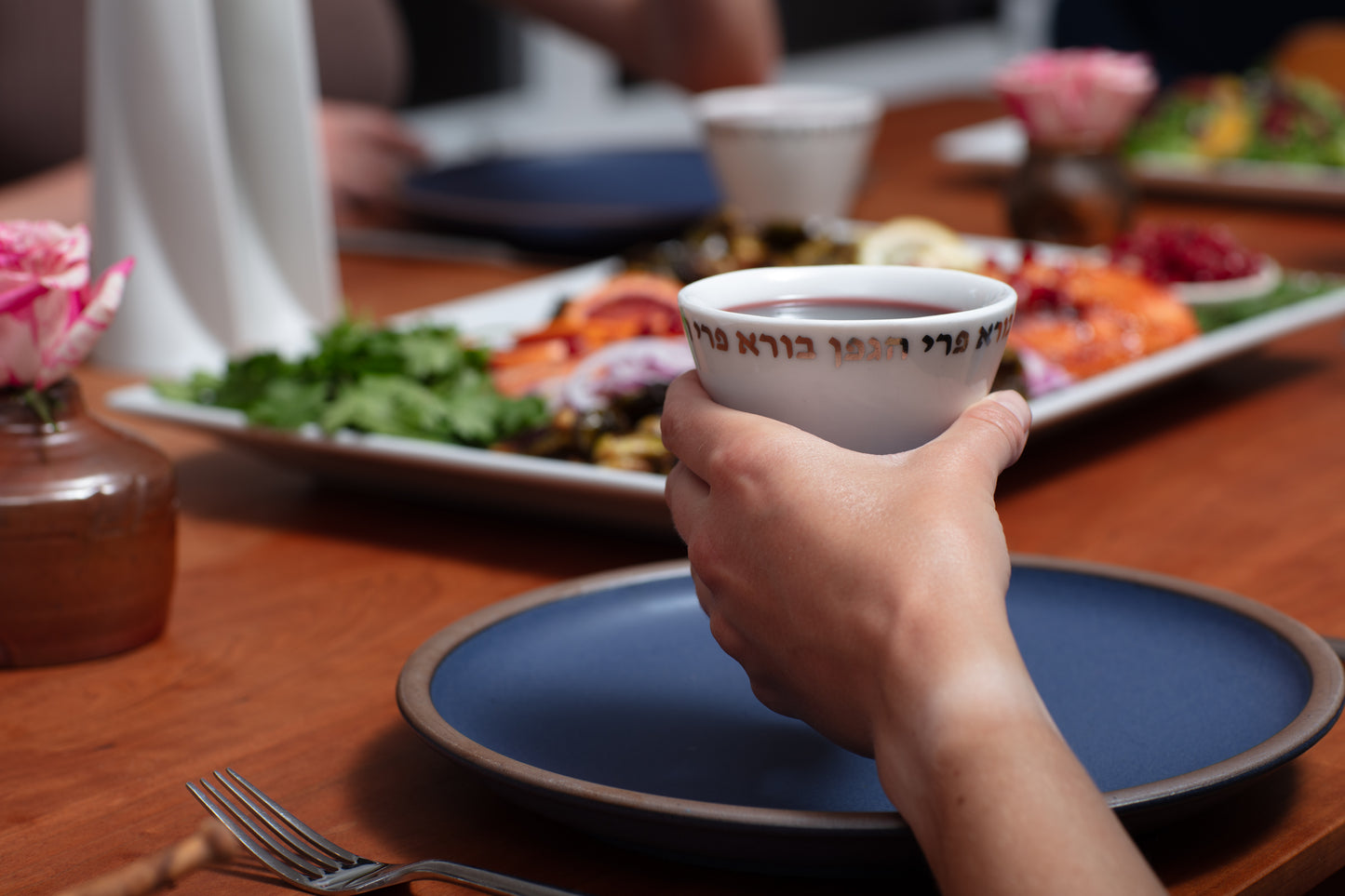 Kiddush cup being held at dinner party, full of wine in front of a set table with food and people