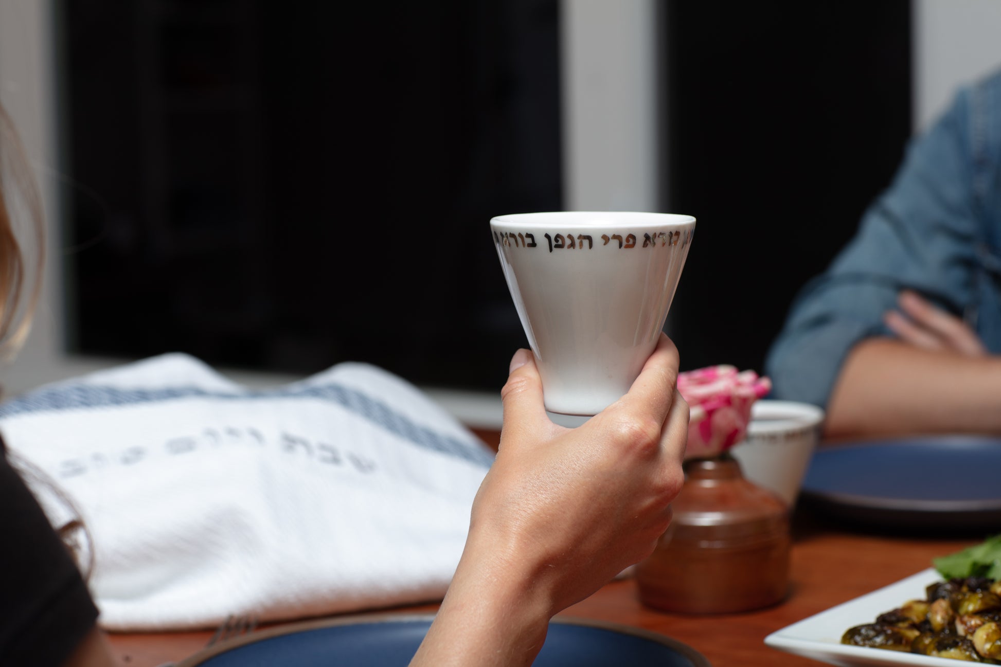 Kiddush cup being held by the base, raised up by a hand at a dinner party, featuring a Challah Cover in the background, available at Green Judaica