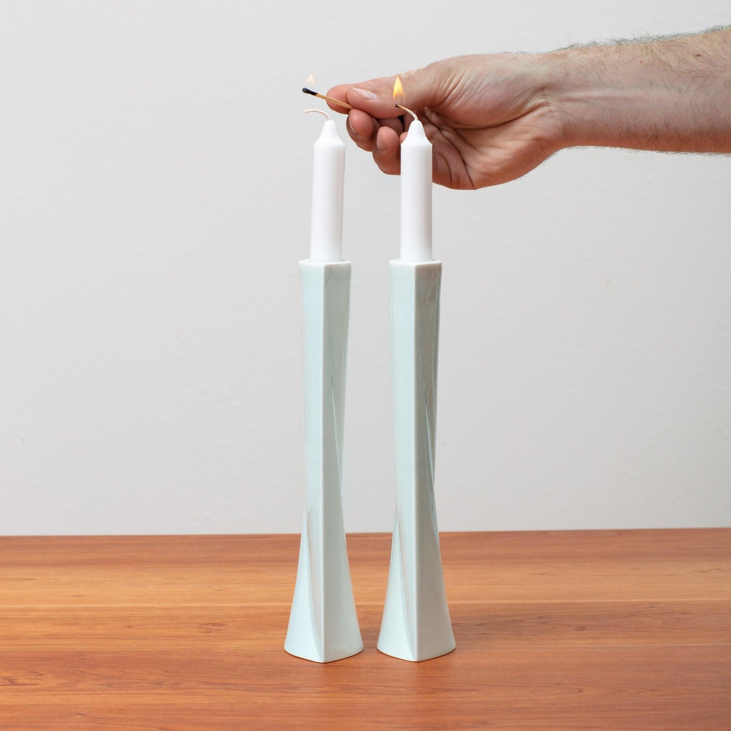 Hand reaching in to light Shabbat Candles placed in white Green Judaica candlesticks with a match